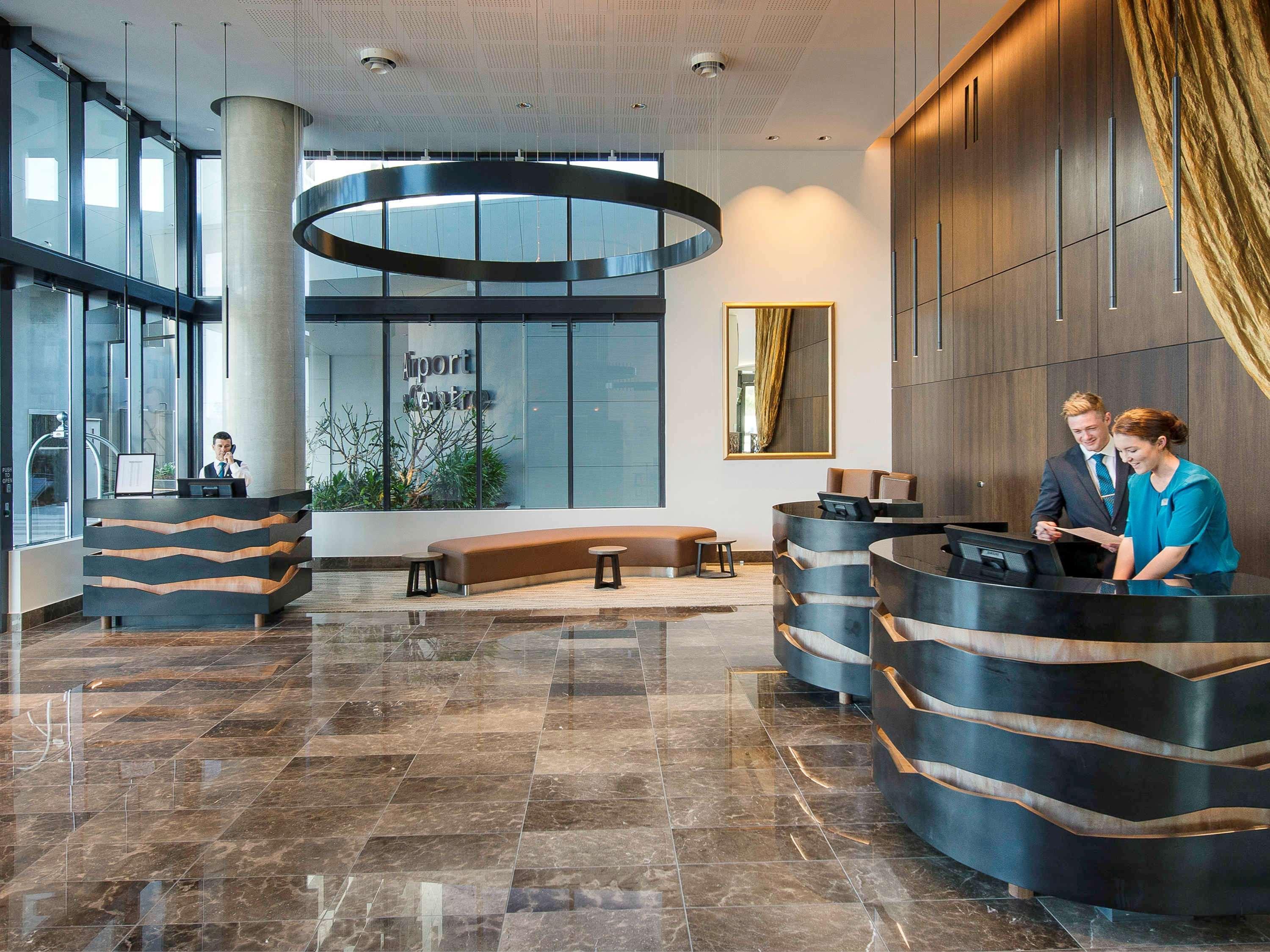 Отель Pullman Brisbane Airport Экстерьер фото The image shows a modern hotel lobby. There are two reception desks made of dark-colored materials, and staff members are attending to guests at these desks. The lobby features large windows that allow natural light to fill the space, and there are p